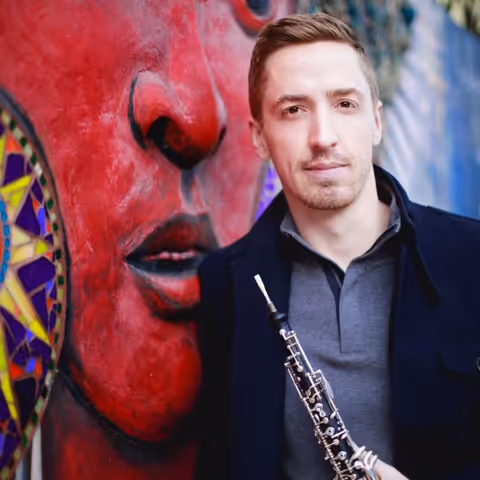 James Button holding an oboe in front of the Tontasin Renance mural in San Francisco, CA
